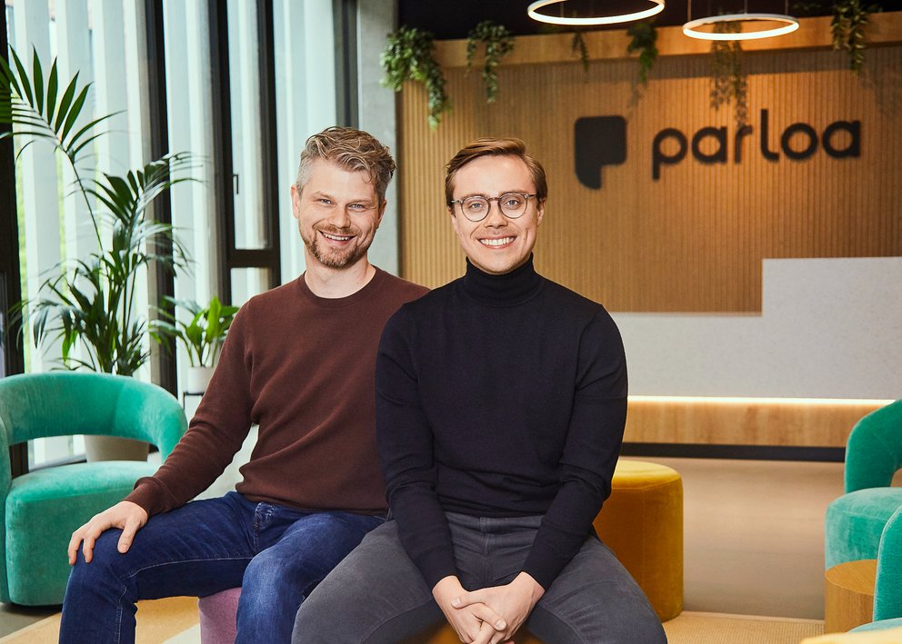 Zwei Männer sitzen nebeneinander in einem modernen Büro mit dem Schriftzug 'parloa' an der Wand im Hintergrund