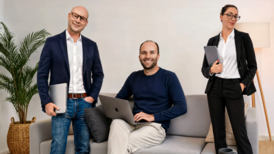In einem hellen Raum steht ein hellgraues Sofa. Auf diesem Sofa sitzt ein Mann mit einem Macbook. Neben dem Sofa stehen ein Mann und eine Frau, die ebenfalls ein Laptop in der Hand halten. 