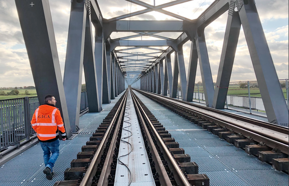 Person mit orangefarbener Warnweste mit Aufschrift 'GIGA' geht auf einer Stahlbrücke entlang der Bahngleise