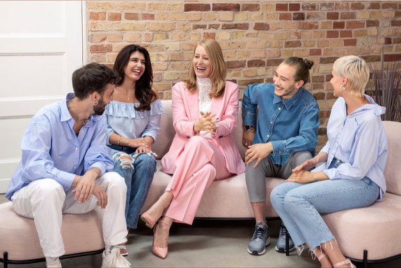 Fünf Personen sitzen in legerer Kleidung auf einem Sofa und Stühlen vor einer Ziegelsteinwand und unterhalten sich.