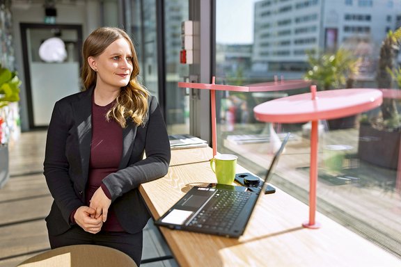 Frau mit langen blonden Haaren, sitzend an einem Holztisch mit Laptop, grüner Tasse und Sonnenbrille, im modernen Büro mit großer Fensterfront