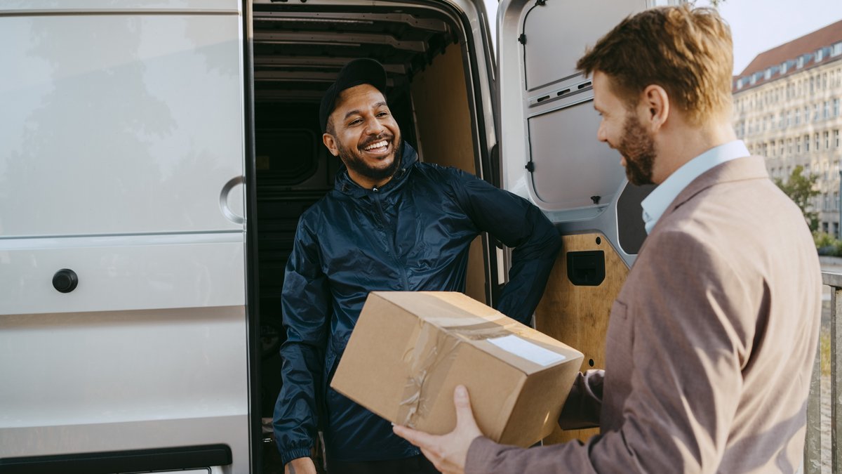 Zwei Männer bei der Übergabe eines Pakets vor einem weißen Lieferwagen mit geöffneter Tür in einer städtischen Umgebung.