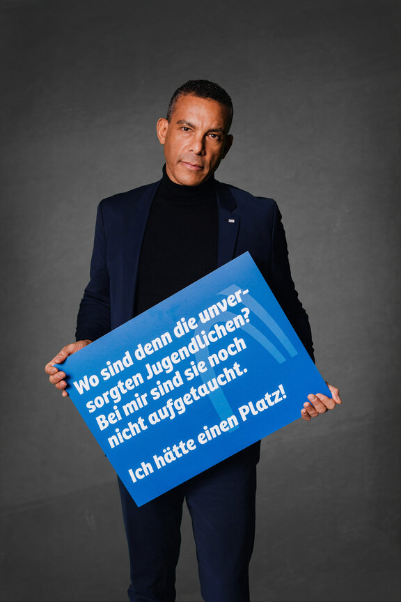 Eine Person steht vor einem neutralen grauen Hintergrund und hält ein großes blaues Schild mit der weißen Aufschrift: "Besser Berufsorientierung statt mehr Bürokratie!"