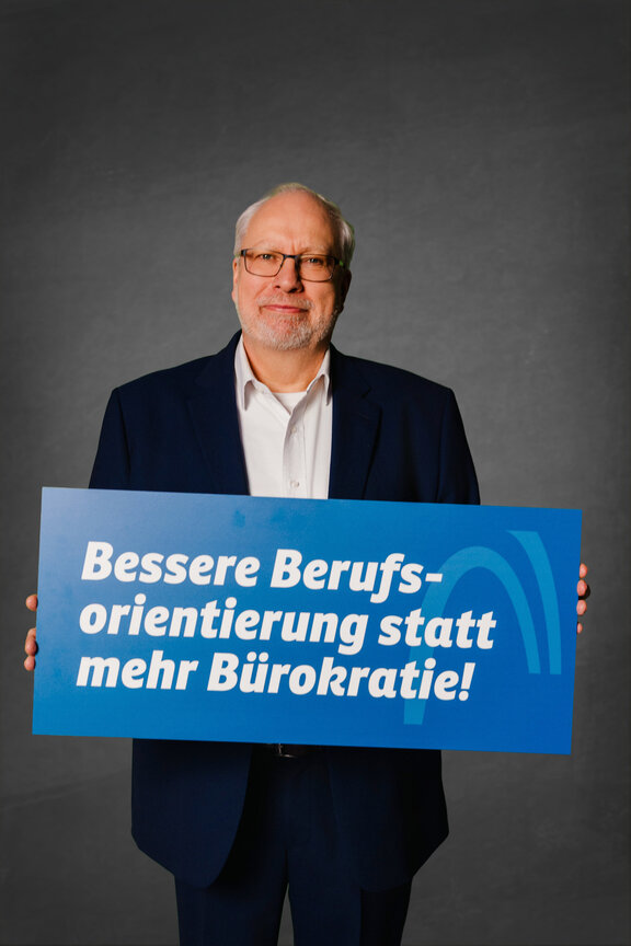 Eine Person steht vor einem neutralen grauen Hintergrund und hält ein großes blaues Schild mit der weißen Aufschrift: "Die Strafabgabe schwächt den Wirtschaftsstandort!"