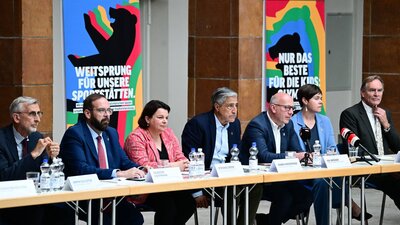 Zu sehen ist ein Langer Tische, an dem mehrere Personen nebeneinander sitzen. Es handelt sich um eine Pressekonferenz.
