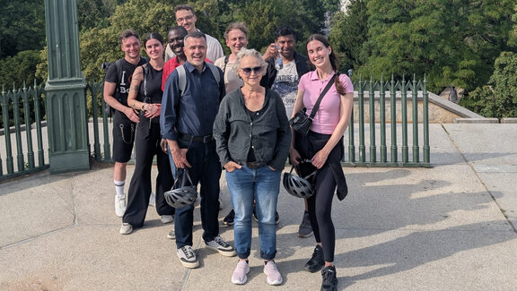 Eine Gruppe von Menschen steht auf einem asphaltiertem Platz. Hinter den Menschen sind mehrere grüne Bäume und Büsche. Das Bild ist aus einer Vogelperspektive fotografiert.