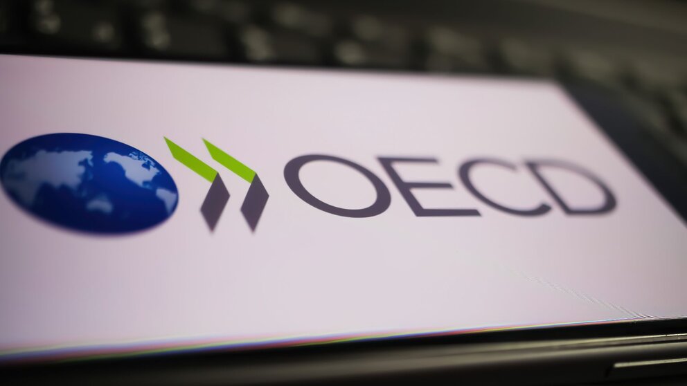 Zu sehen ist ein Smartphone, welches auf einer schwarzen PC-Tastatur liegt. Auf dem Bildschirm des Handys ist das Logo der OECD zu sehen.