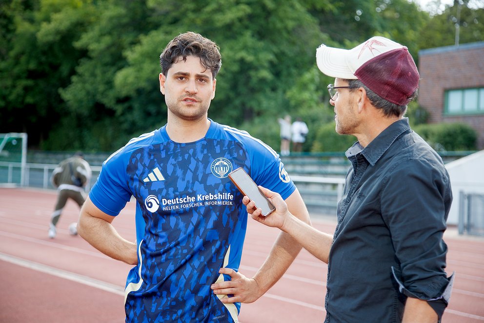 Junger Mann im blauen Sporttrikot mit 'Deutsche Krebshilfe' Logo wird auf einer Laufbahn von einem Mann mit Baseballkappe interviewt