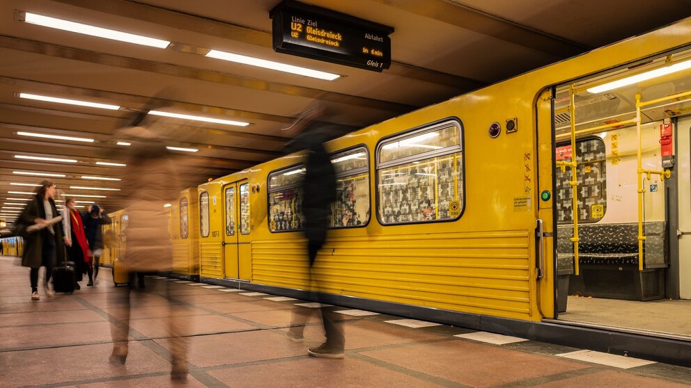 Man sieht eine wartende U-Bahn in Berlin an einer Haltestelle, während Menschen neben der Bahn laufen.