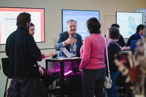 Zu sehen ist ein älterer Herr, der hinter einem Stand sitzt. Hinter ihm befinden sich mehrere Bildschirme. Vor dem Stand stehen mehrere Personen, die sich mit dem älteren Herren unterhalten. Sie befinden sich auf der Digital-Konferenz der IHK Berlin und HTW Berlin.
