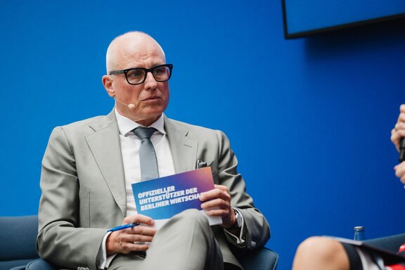 Zu sehen ist ein Herr im grauen Anzug mit Glatze und Brille. Er hält eine Moderationskarte in der Hand.