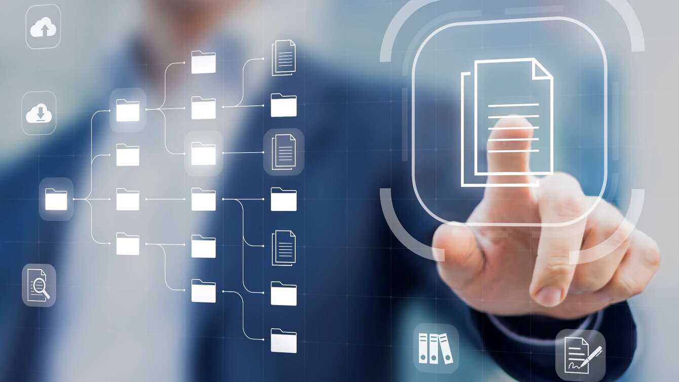 Zu sehen ist eine Hand, die auf ein technisches Symbol zeigt, welches wie ein Hologramm im Bild schwebt. Im Hintergrund ist unscharf ein Mann im Anzug zu erkennen, zu dem die Hand gehört. Es handelt sich um ein Symbolbild zur Digitalisierung.