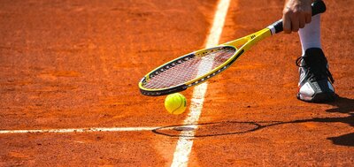 Zu sehen ist ein gelber Tennisball und darüber ein Tennisschläger und ein Fuß.