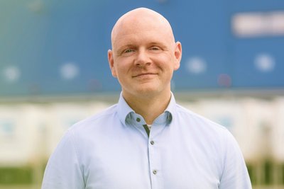 Mann in hellblauem Hemd und grauer Hose steht vor einem Logistikzentrum mit Laderampen und Containern.