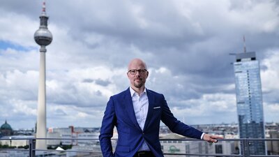 Zu sehen ist ein Mann im Anzug, der auf einem Balkon steht. Im Hintergrund ist die Stadt Berlin zu sehen.