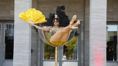 Performerin in glitzerndem Kostüm mit schwarzem Federkopfschmuck und goldenem Fächer sitzt in einem übergroßen Sektglas vor dem Eingang der Berliner Messehallen.