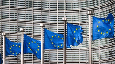 Zu sehen ist eine Hausfassade im Hintergrund, im Vordergrund wehen mehrere an Masten befestigte EU-Flaggen im Wind.