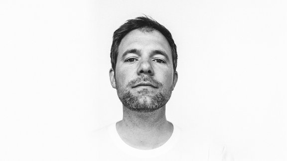 Mann mit kurzem dunklem Haar, trägt ein weißes T-Shirt, vor weißem Hintergrund