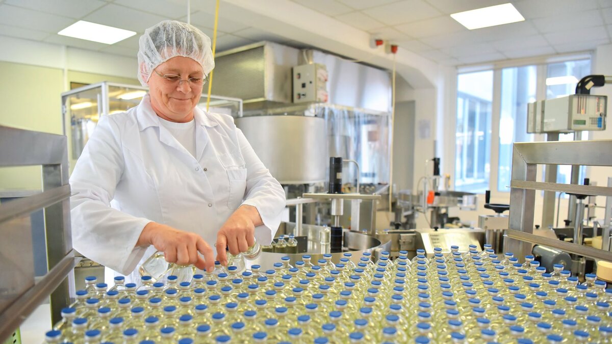 Eine Labormitarbeiterin mit weißem Kittel und Haarnetz nimmt Medikamentenfläschchen von einem Fließband.
