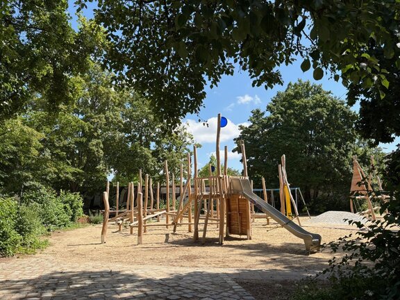 Auf dem Bild ist ein Spielplatz zusehen, um ihn herum stehen Bäume und Büsche die Grün sind. Der Himmel ist Blau und ein paar Wolken sind zusehen. 