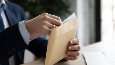 Zu sehen ist die Hände einer Person im Anzug, welche an einem Schreibtisch vor einem aufgeklappten Laptop sitzt und einen Brief in einen braunen Umschlag packt.