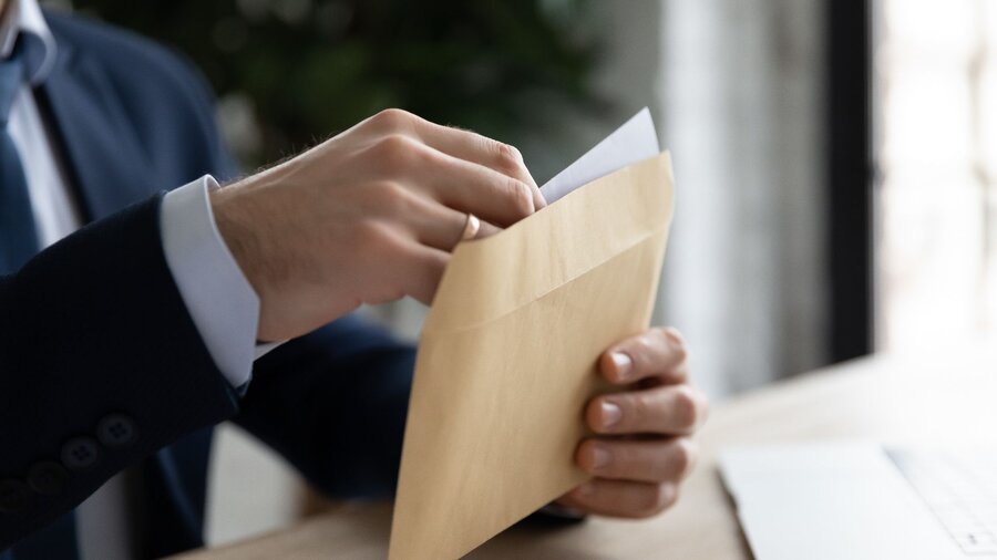 Zu sehen ist die Hände einer Person im Anzug, welche an einem Schreibtisch vor einem aufgeklappten Laptop sitzt und einen Brief in einen braunen Umschlag packt.