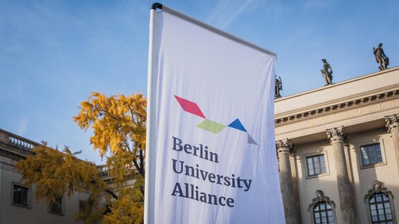 Weiße Flagge mit der Aufschrift "Berlin University Alliance" vor einem alten gebäude mit einem herbstlichen Baum.