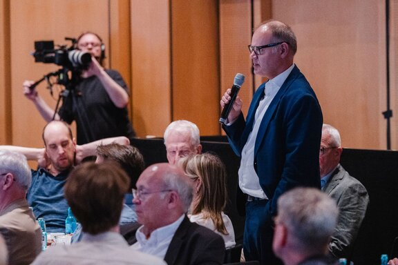 Zu sehen ist ein älterer Herr im Anzug, welcher in ein Mikrofon spricht. Er ist Teil des Publikums, im Hintergrund steht eine Person, die die Szene mit einer Filmkamera aufnimmt.