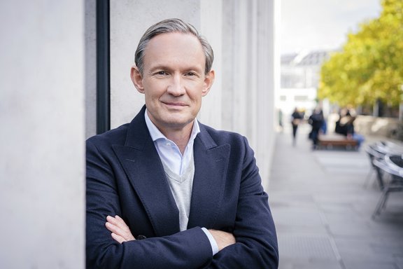 Mann mit grauem Haar, dunkelblauem Mantel, hellblauem Hemd und grauem Pullover lehnt mit verschränkten Armen an einer weißen Wand im Freien.