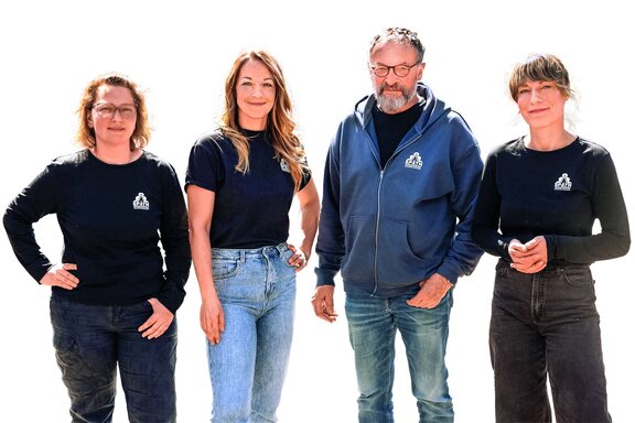 Zu sehen sind vier Personen, drei Frauen und ein älterer Mann. Sie schauen freundlich in die Kamera und stehen vor einem neutral-weißem Hintergrund.