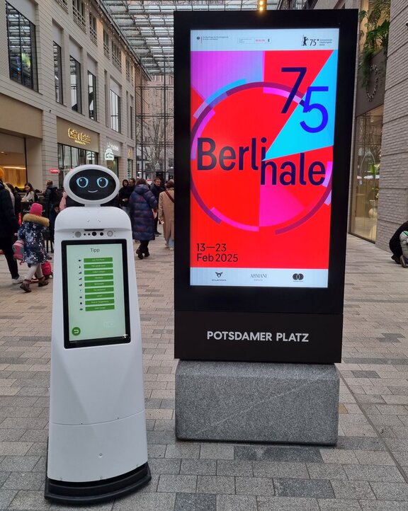 Ein humanoider Roboter steht neben einem Werbedisplay der Berlinale 2025. Im Hintergrund ist eine Einkaufspassage zu sehen. 