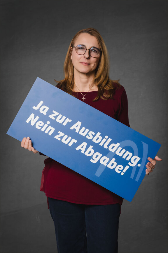 Eine Person steht vor einem neutralen grauen Hintergrund und hält ein großes blaues Schild mit der weißen Aufschrift: "Ja zu Ausbildung. Nein zur Abagabe!""