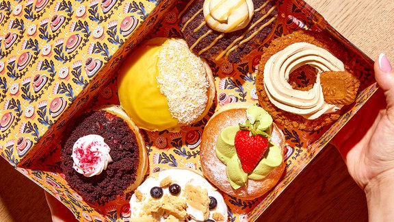 Hand hält eine bunte Schachtel mit sechs verschiedenen Mini-Törtchen, darunter mit Erdbeere, Schokolade, Vanillecreme und Kokosraspeln