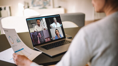 Zu sehen ist eine Frau in weißem Oberteil, welcher man von hinten über die Schulter schaut. Sie sitzt vor einem Laptop und nimmt an einem Videocall teil, während sie ein Blatt in einer Hand hält.