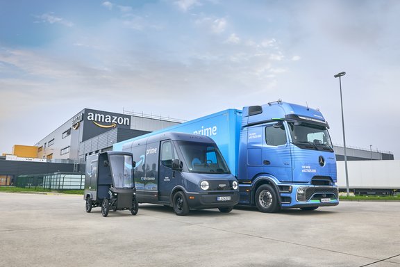 Drei blaue Amazon Prime Lieferfahrzeuge unterschiedlicher Größe vor einem Amazon-Logistikzentrum mit sichtbarem Amazon-Logo.