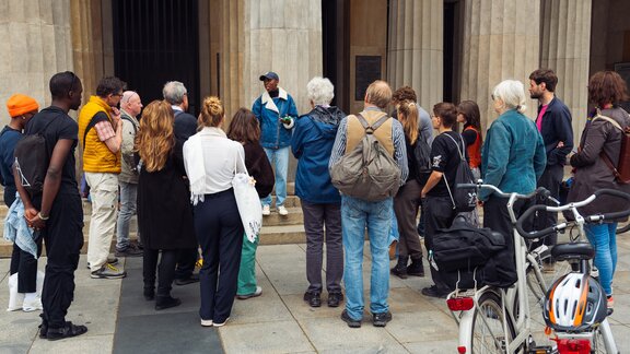 Zu sehen ist eine Gruppe Menschen die vor einem großen Gebäude mit Säulen stehen. Eine Person redet zu ihnen. Es ist eine Stadtführung und die Gruppe befindet sich am Brandenburger Tor.