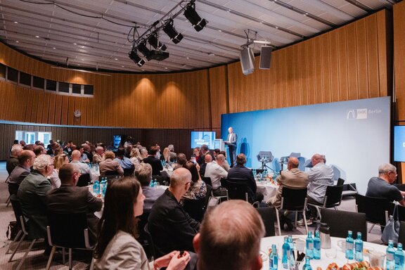 Zu sehen ist ein großer Saal, in welchem viel Publikum sitzt. Dieses schaut auf eine Bühne, auf der eine Person am Redepult steht.