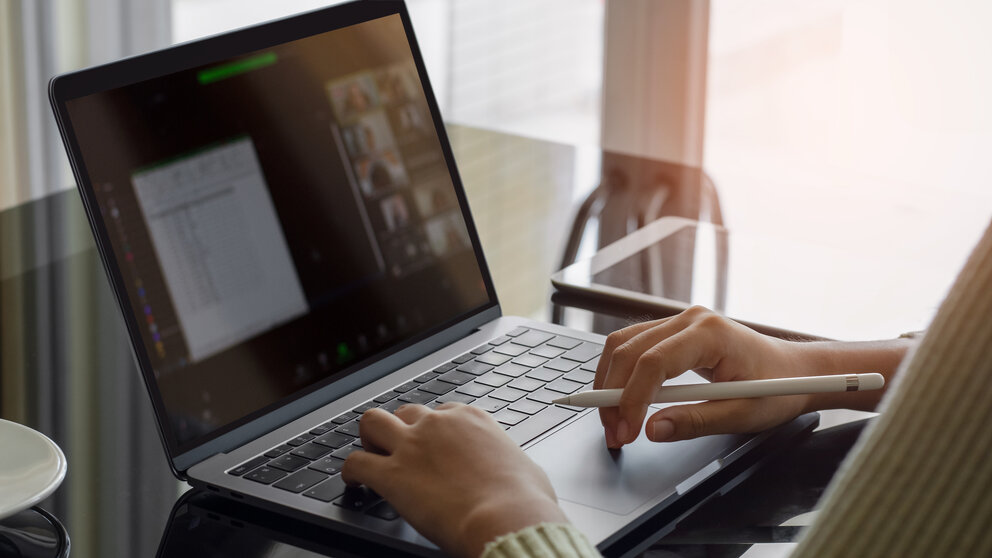Hände einer Person, die an einem Laptop arbeitet. Auf dem Bildschirm ist unscharf ein Videokonferenzfenster zu erkennen. Die Person hält einen Eingabestift und sitzt an einem Tisch in einem hellen Raum.