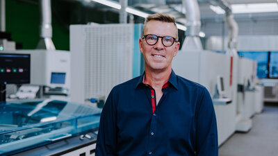 Ein Mann mit dunkler Kleidung und Brille steht vor digitalen Druckmaschinen.