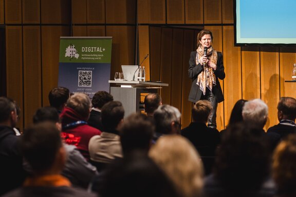 Zu sehen ist Publikum von hinten. Über den Köpfen sieht man eine Bühne auf der eine Person steht und in ein Mikrofon spricht.