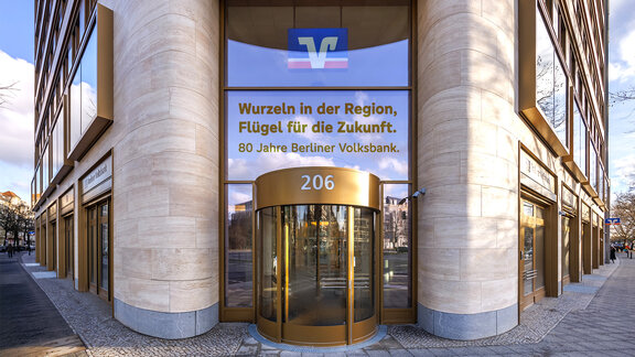 Zu sehen ist der Eingang einer Bankfiliale. Eine Glasfront wird von zwei Stein-Säulen umschlossen, im Zentrum befindet sich eine gläserne Drehtür. Es handelt sich um eine Filiale der Berliner Volksbank in Berlin.