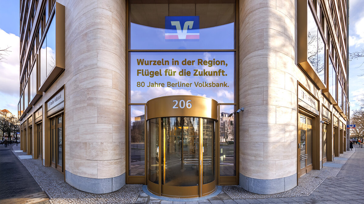 Zu sehen ist der Eingang einer Bankfiliale. Eine Glasfront wird von zwei Stein-Säulen umschlossen, im Zentrum befindet sich eine gläserne Drehtür. Es handelt sich um eine Filiale der Berliner Volksbank in Berlin.