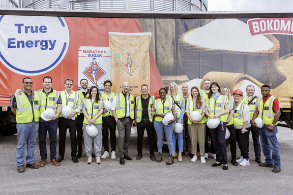 Eine größere Gruppe von Personen in Warnwesten steht für ein Gruppenfoto vor einem Lastwagen mit Logos von ‚True Energy‘ und der Firma Bokomo. Einige halten Schutzhelme in der Hand.