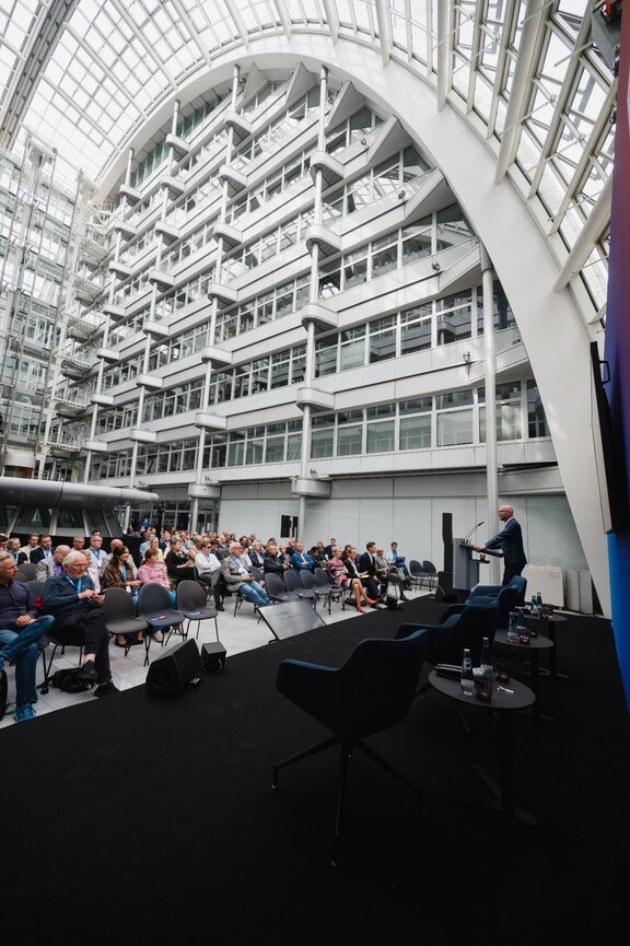 Zu sehen ist ein großes Atrium. Man schaut von einer Bühne, auf der ein Sprecher steht, auf ein bestuhltes Publikum.