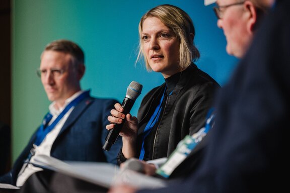 Drei sitzende Speaker auf einer Bühne. In der Mitte eine Frau mit Mikrofon.