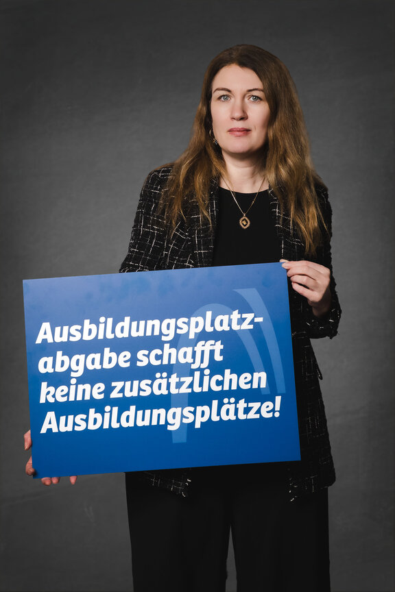 Eine Person steht vor einem neutralen grauen Hintergrund und hält ein großes blaues Schild mit der weißen Aufschrift: "Ausbildungsplatzabgabe schafft keine zusätzlichen Ausbildungsplätze!"