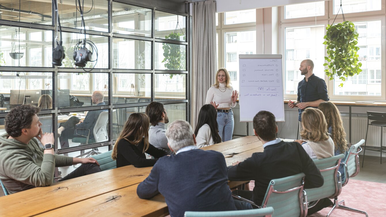 Zu sehen ist ein Geschäftsmeeting, bei dem mehrere Personen an einem Tisch sitzen und zwei Vortragenden zuhören, die vor einem Whiteboard stehen.