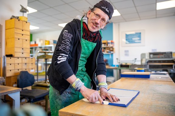 Person mit grünem Schürzenkleid und schwarzer Kapuzenjacke arbeitet an einem Tisch in einer Buchbinderei mit Werkzeugen und Regalen im Hintergrund.