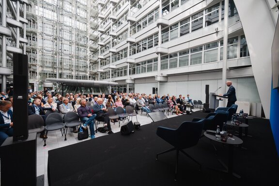 Zu sehen ist Publikum vom Blick der Bühne aus. Die Personen sitzen in Stuhlreihen in einem großen Atrium.