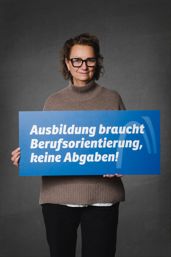 Eine Person steht vor einem neutralen grauen Hintergrund und hält ein großes blaues Schild mit der weißen Aufschrift: "Ja zur Ausbildung. Nein zur Abgabe!"